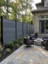 Light gray composite fence lining a stone patio with outdoor chairs and a round fire pit